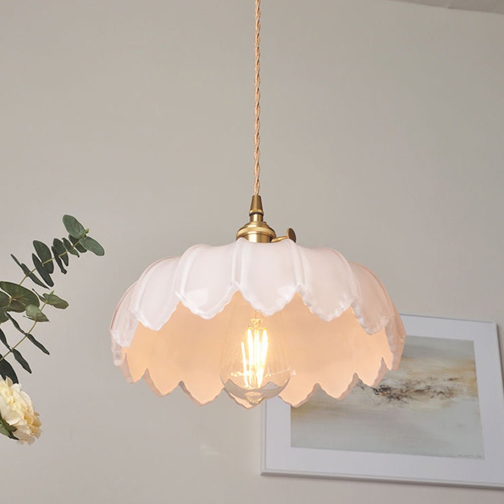 Een stijlvolle plafondlamp met een lichtroze glazen kap in de vorm van bloemblaadjes, die een warme en uitnodigende sfeer creëert. De lamp heeft een gouden fitting en een decoratieve, gevlochten kabel, die bijdraagt aan het moderne ontwerp. Op de achtergrond is een subtiele kunstafdruk te zien, samen met een groene plant die de uitstraling van de ruimte verfraait.